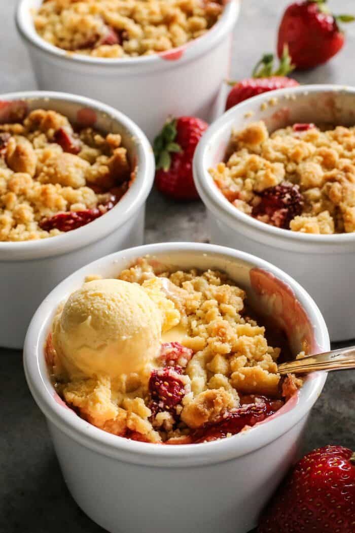 four white ramekins of strawberry rhubarb crumble, one with a scoop of ice cream and a spoon