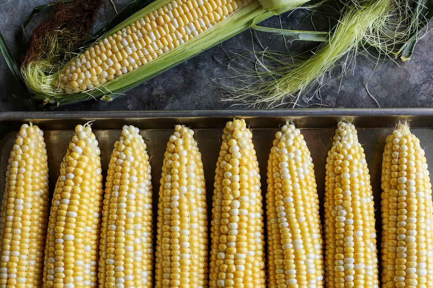 a pan of husked sweet corn plus an ear of corn that is halfway husked