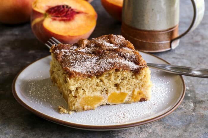 a slice of peach cake on a plate, with sliced peaches and a coffee cup in the background