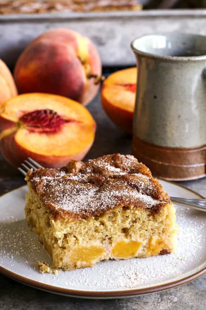 a piece of peach cake on a plate, plus fresh peaches and a cup of coffee