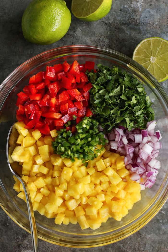 a large clear bowl with diced pineapple, red pepper, red onion, jalapeno, and cilantro, plus fresh limes