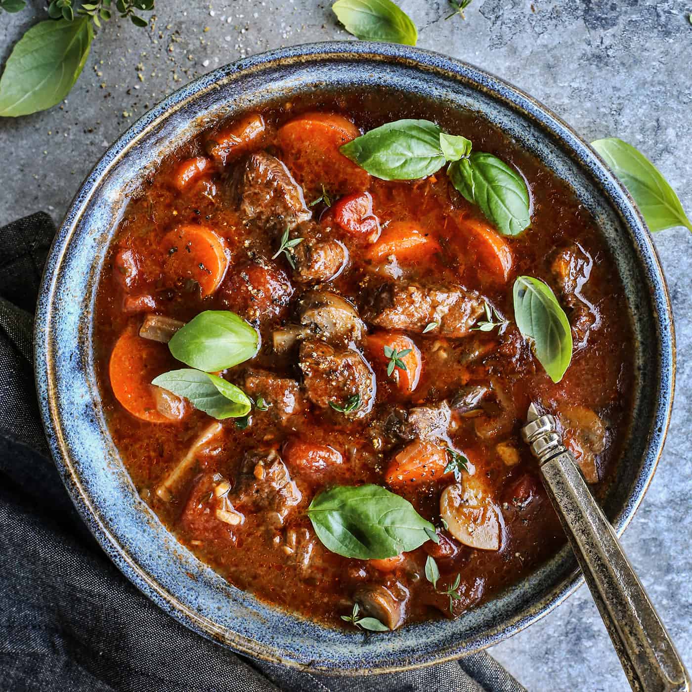 This cozy Italian beef stew is an easy slow cooker recipe with juicy bites of beef and veggies in a rich, herby tomato broth with red wine. a blue pottery bowl with Italian beef soup, garnished with fresh basil leaves