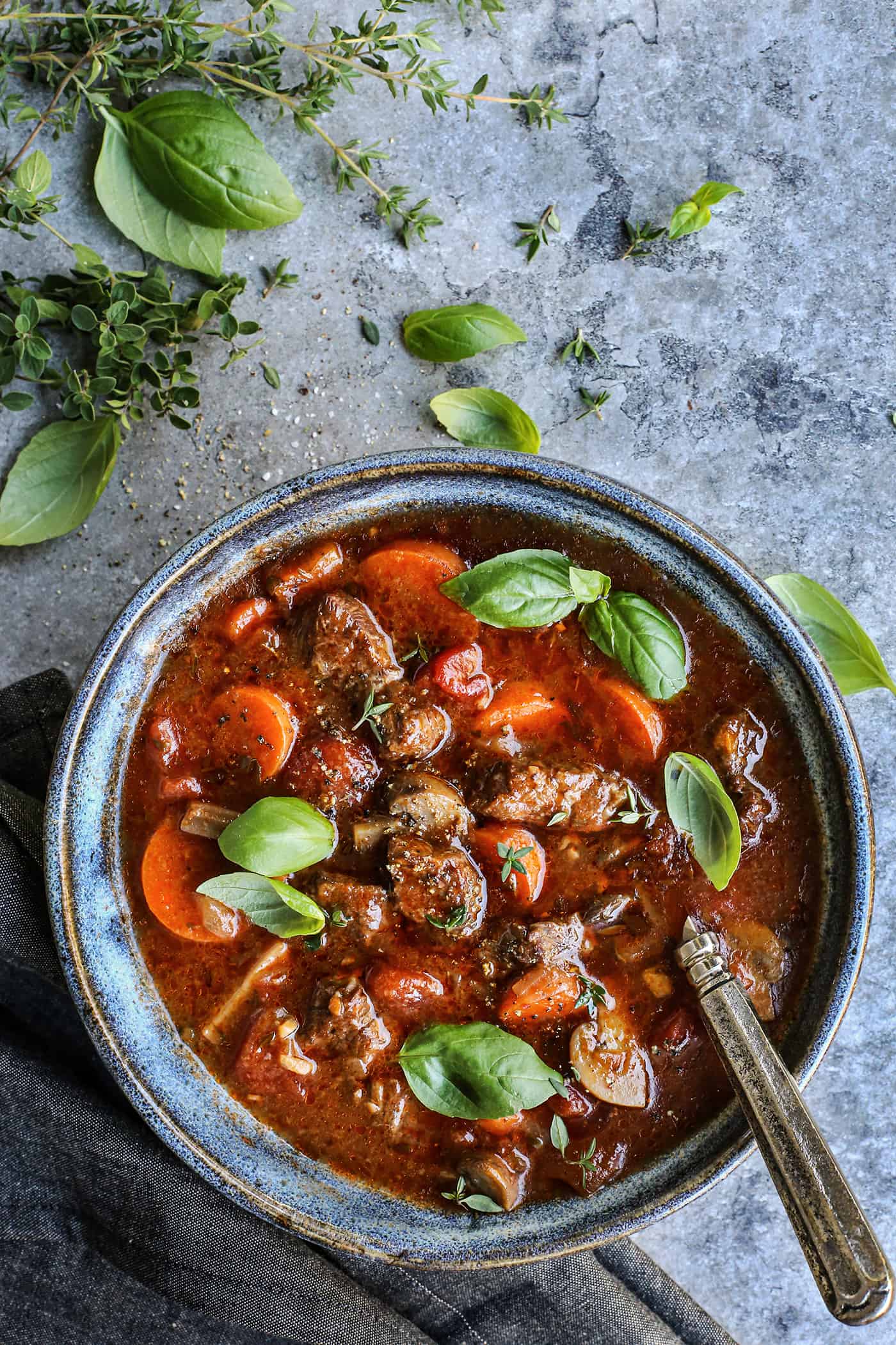 blue pottery bowl of Italian beef stew, plus fresh thyme, basil, and oregano