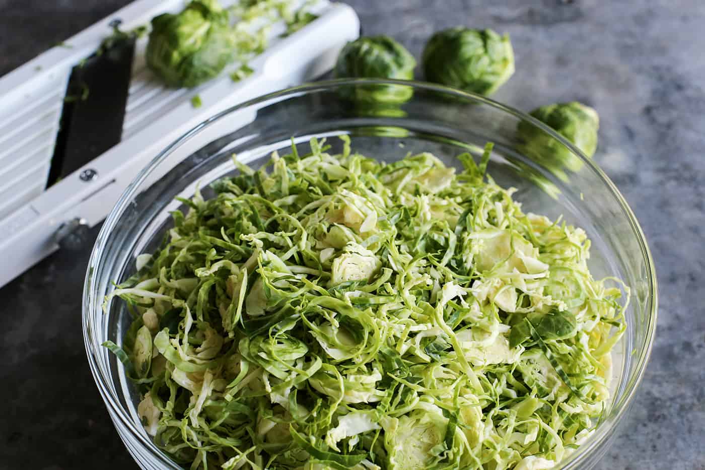 Shaved Brussels sprouts in a clear bowl, a mandoline cutter in the background.