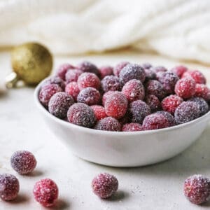 Sugared cranberries in a small bowl next to scattered cranberries and Christmas ornaments.