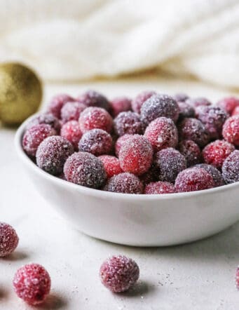 Sugared cranberries in a small bowl next to scattered cranberries and Christmas ornaments.