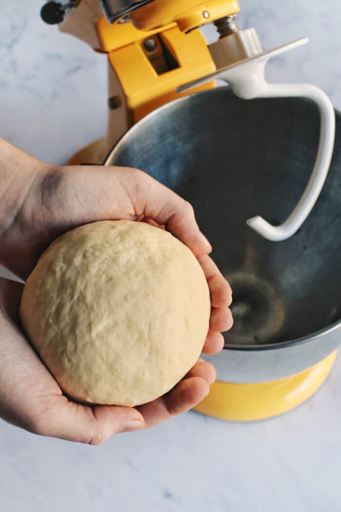 a yellow stand mixer fitted with a dough hook and two hands holding a ball of pizza dough