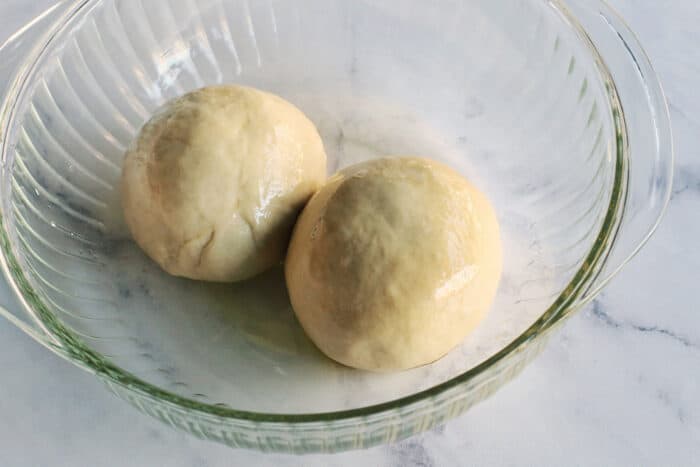 two balls of pizza dough rolled in olive oil, in a clear bowl