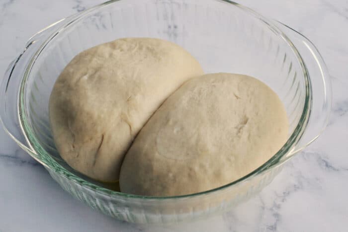 two balls of pizza dough rising in a clear bowl