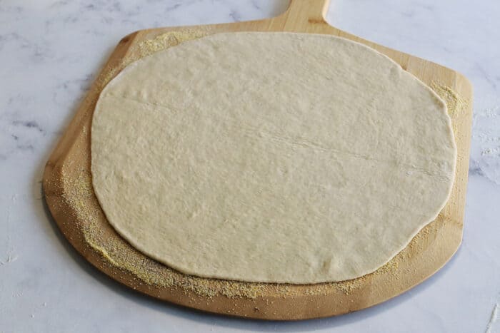 pizza dough rolled into a large circle, on top of a wood pizza peel with cornmeal