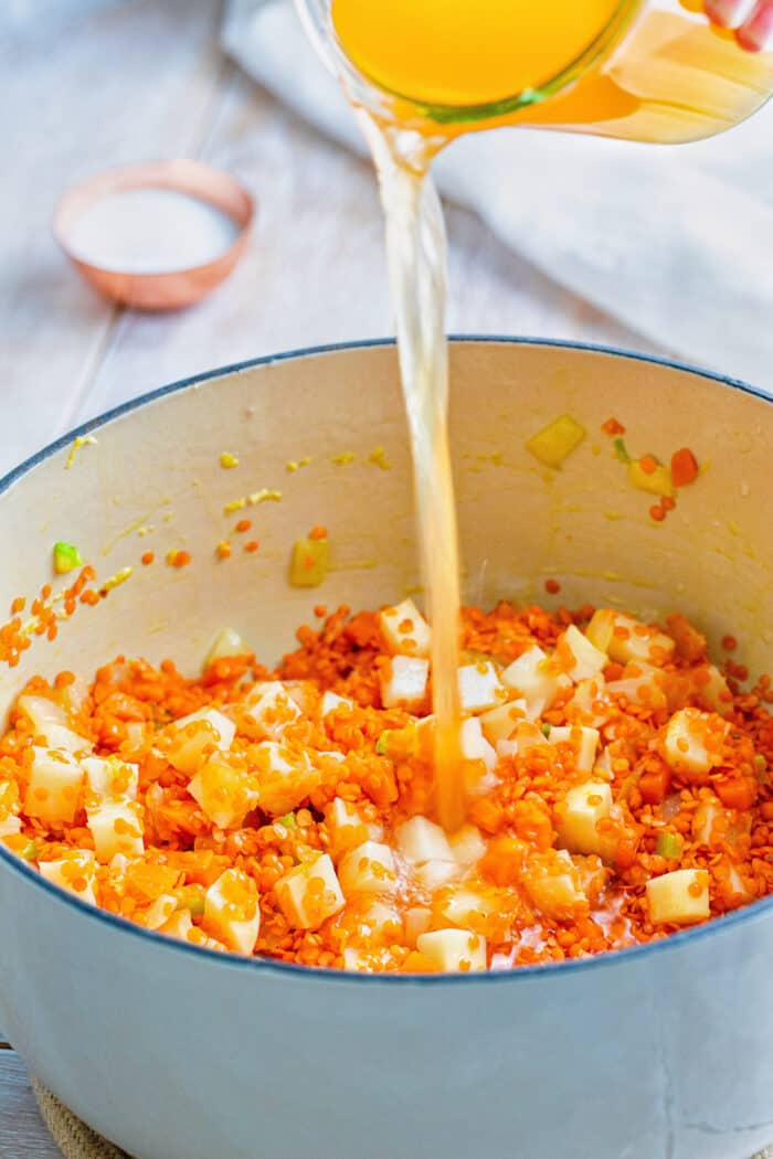 pouring stock into a white pot of red lentils and diced potato