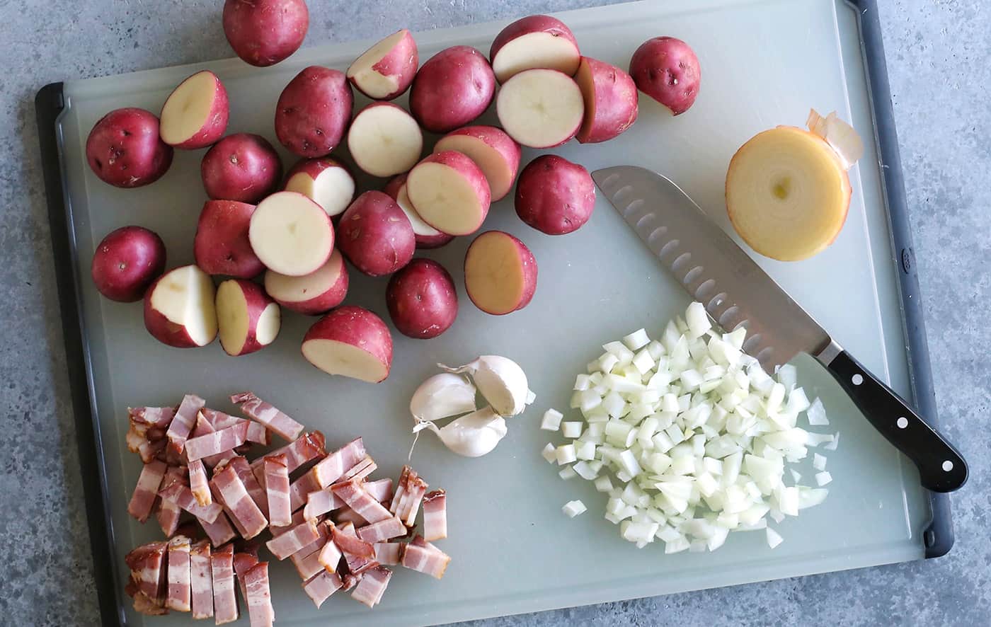 potatoes, onion, garlic, and bacon on a cutting board with a chef's knife