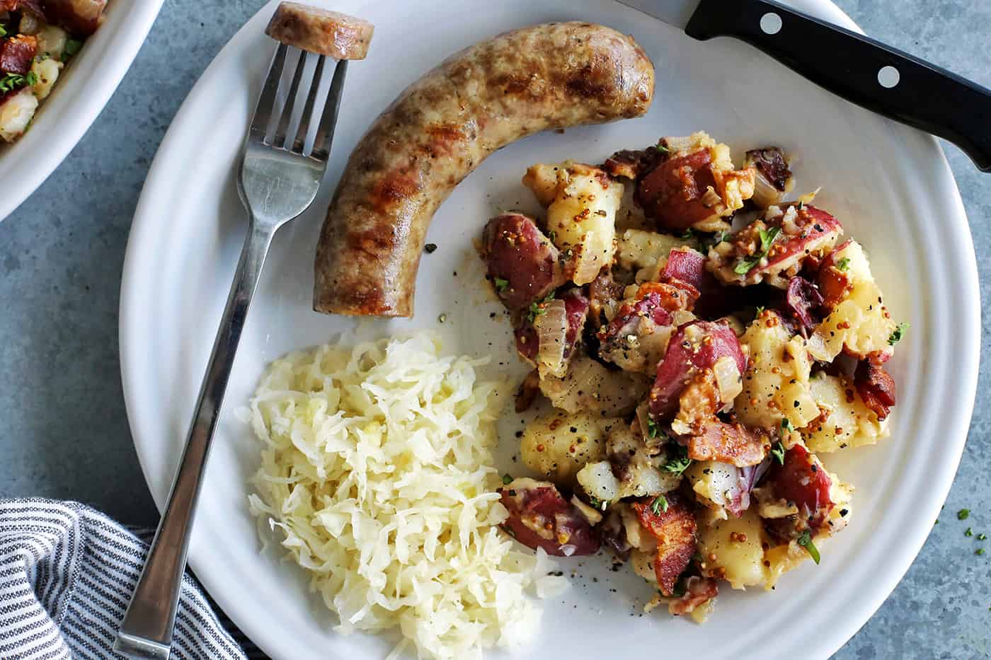 a white plate with German potato salad, sauerkraut, and a bratwurst