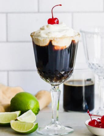 Mexican coffee cocktail in a stemmed glass, plus fresh limes and stemmed maraschino cherries