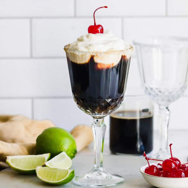 Mexican coffee cocktail in a stemmed glass, plus fresh limes and stemmed maraschino cherries