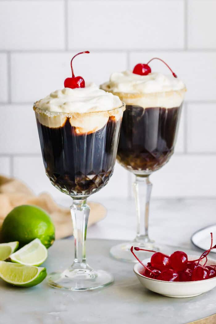 two glasses of Mexican coffee in stemmed glasses, with fresh limes and maraschino cherries