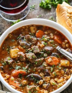 a white bowl of beef barley stew, plus crusty bread and a glass of red wine