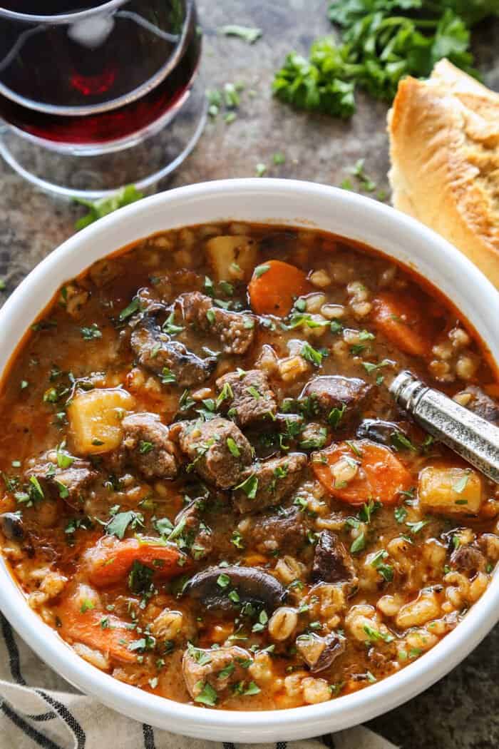 a white bowl of beef barley stew, plus crusty bread and a glass of red wine