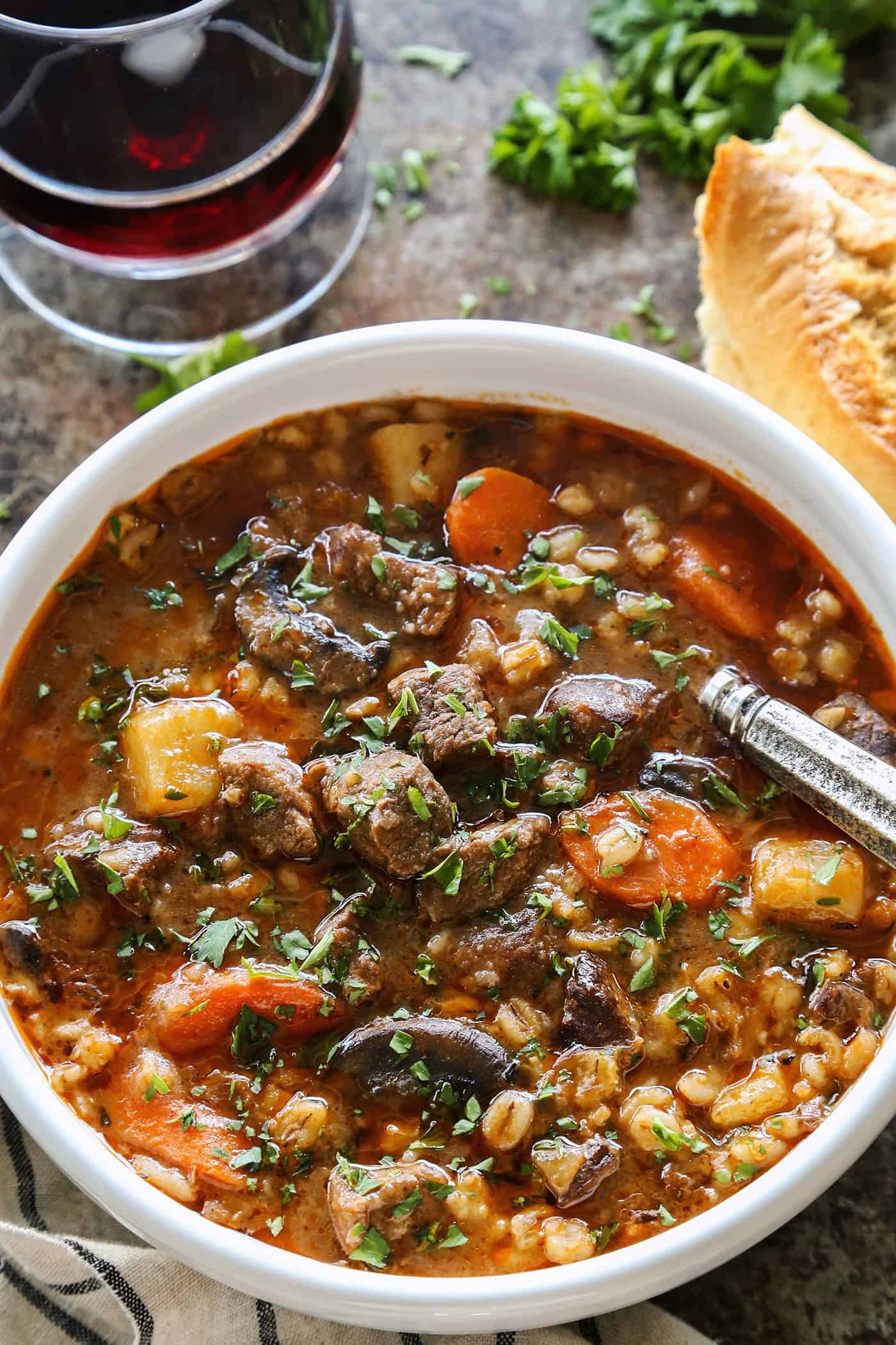 a white bowl of beef barley soup, plus crusty bread and a glass of red wine