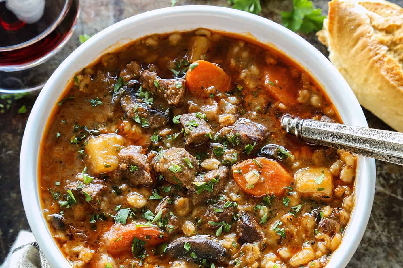 beef barley soup in a white bowl, plus crusty bread and glass of red wine