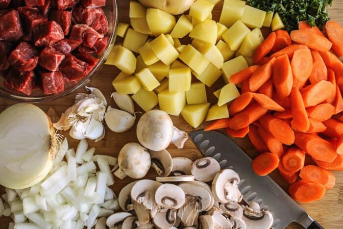 ingredients being cut on a wood cutting board: beef, potatoes, carrots, mushrooms, garlic, and onion