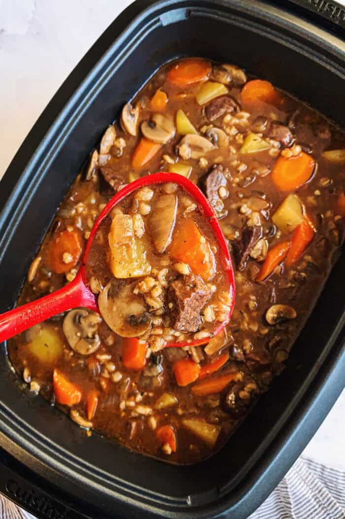 a red ladle of beef stew, over a slow cooker full of beef stew