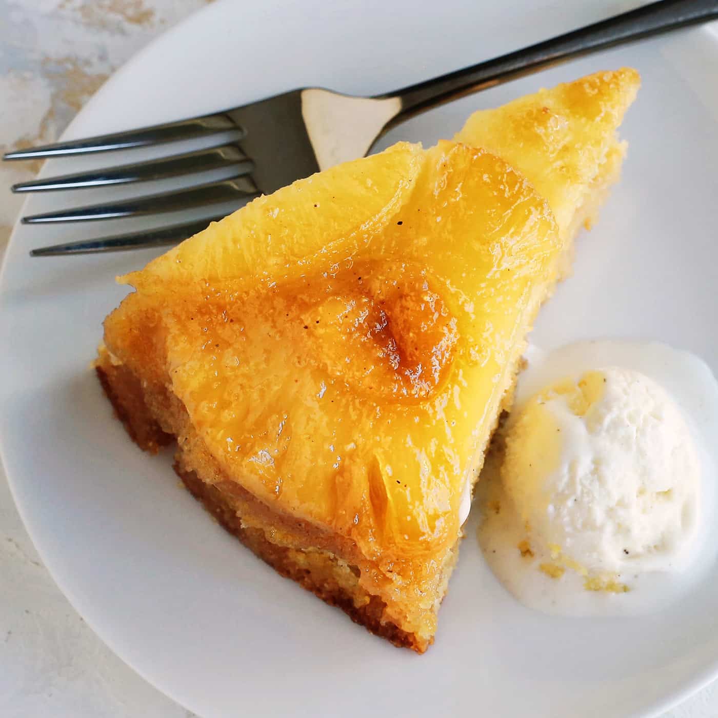 This classic pineapple upside-down cake recipe has a rich, buttery cake with cozy spices and a caramelized brown sugar pineapple topping! Close up of a slice of homemade pineapple upside down cake next to a scoop of ice cream on a plate, with a fork.