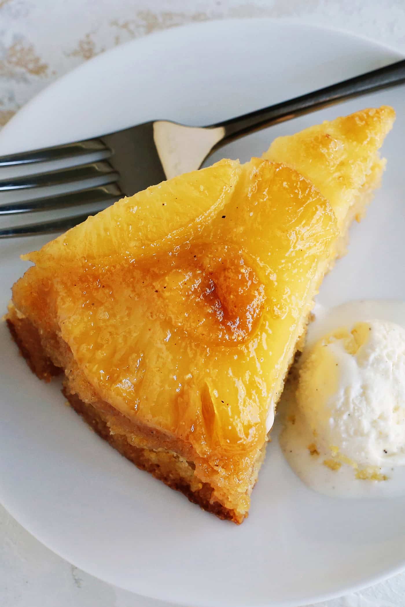 This classic pineapple upside-down cake recipe has a rich, buttery cake with cozy spices and a caramelized brown sugar pineapple topping! Close up of a slice of homemade pineapple upside down cake next to a scoop of ice cream on a plate, with a fork.