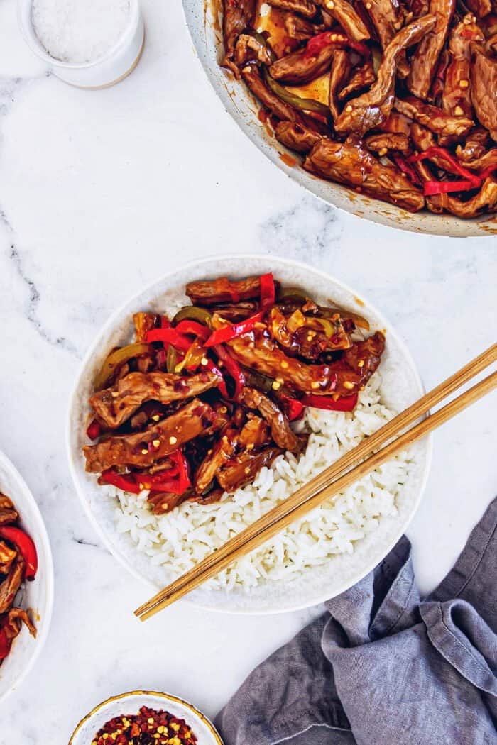 a serving of Beijing beef over white rice in a white bowl, with wood chopsticks