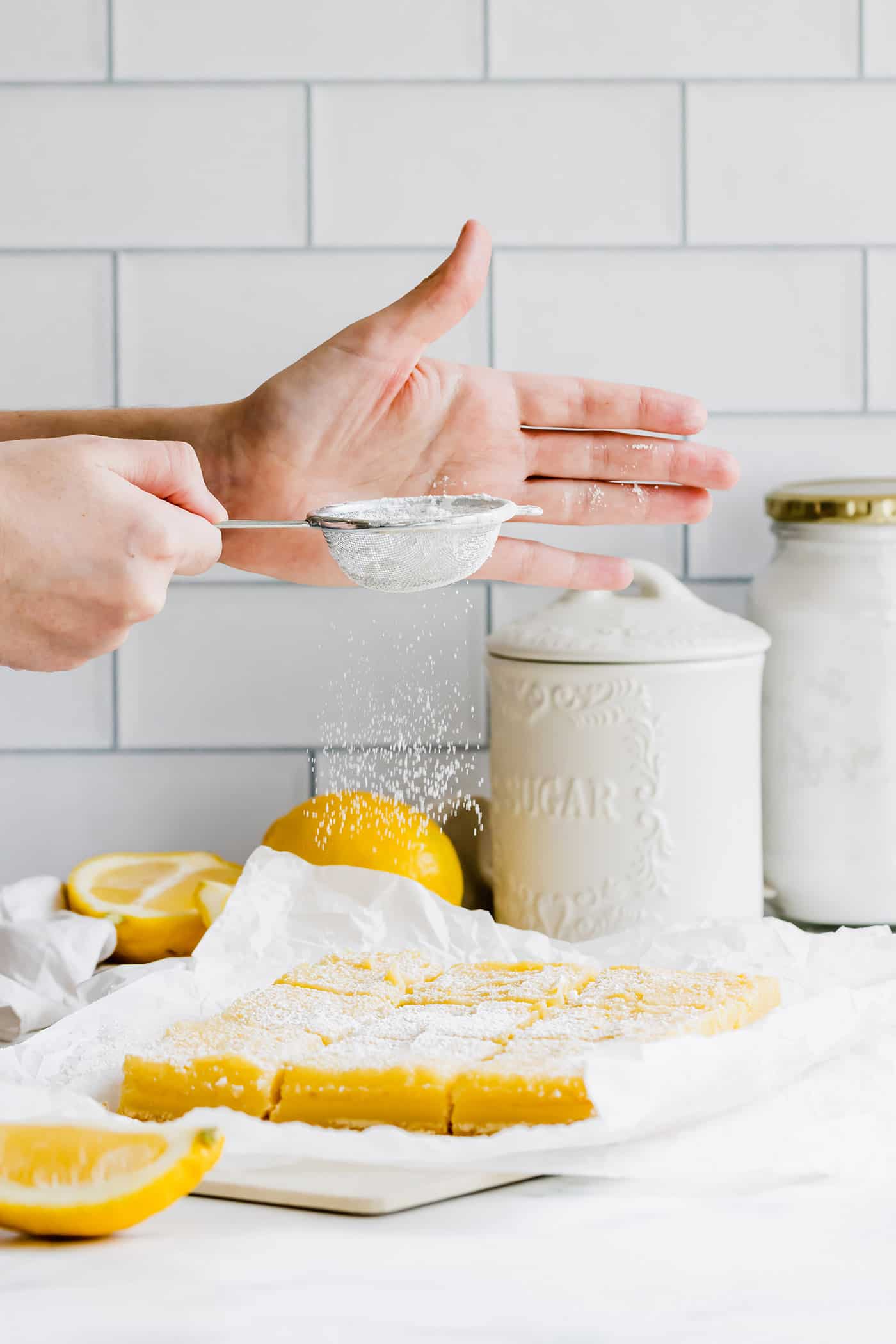 hands sprinkling powdered sugar over cut lemon bars on a piece of parchment