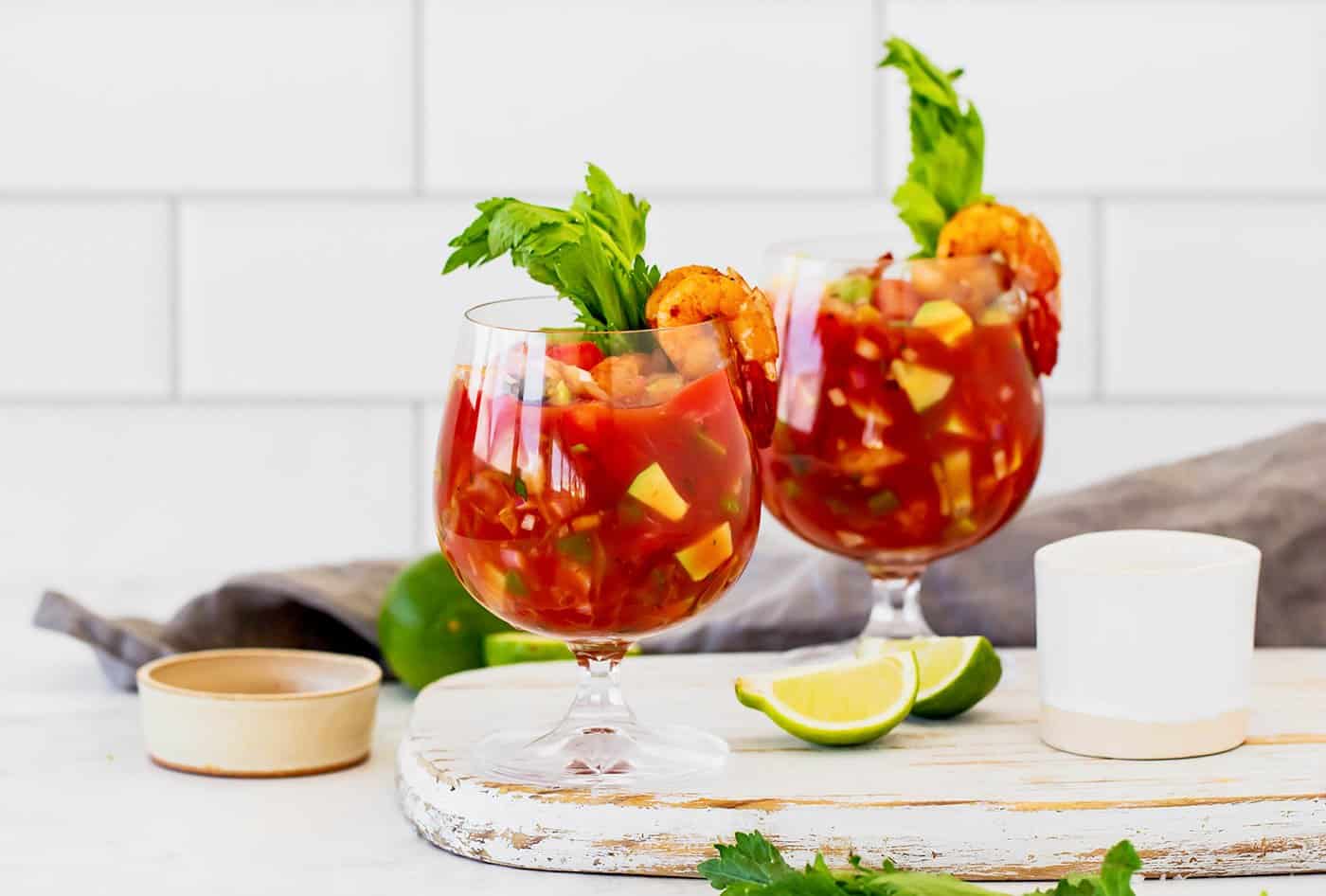 a white cutting board with two stemmed glasses of an appetizer made with tomato juice, lime, and shrimp