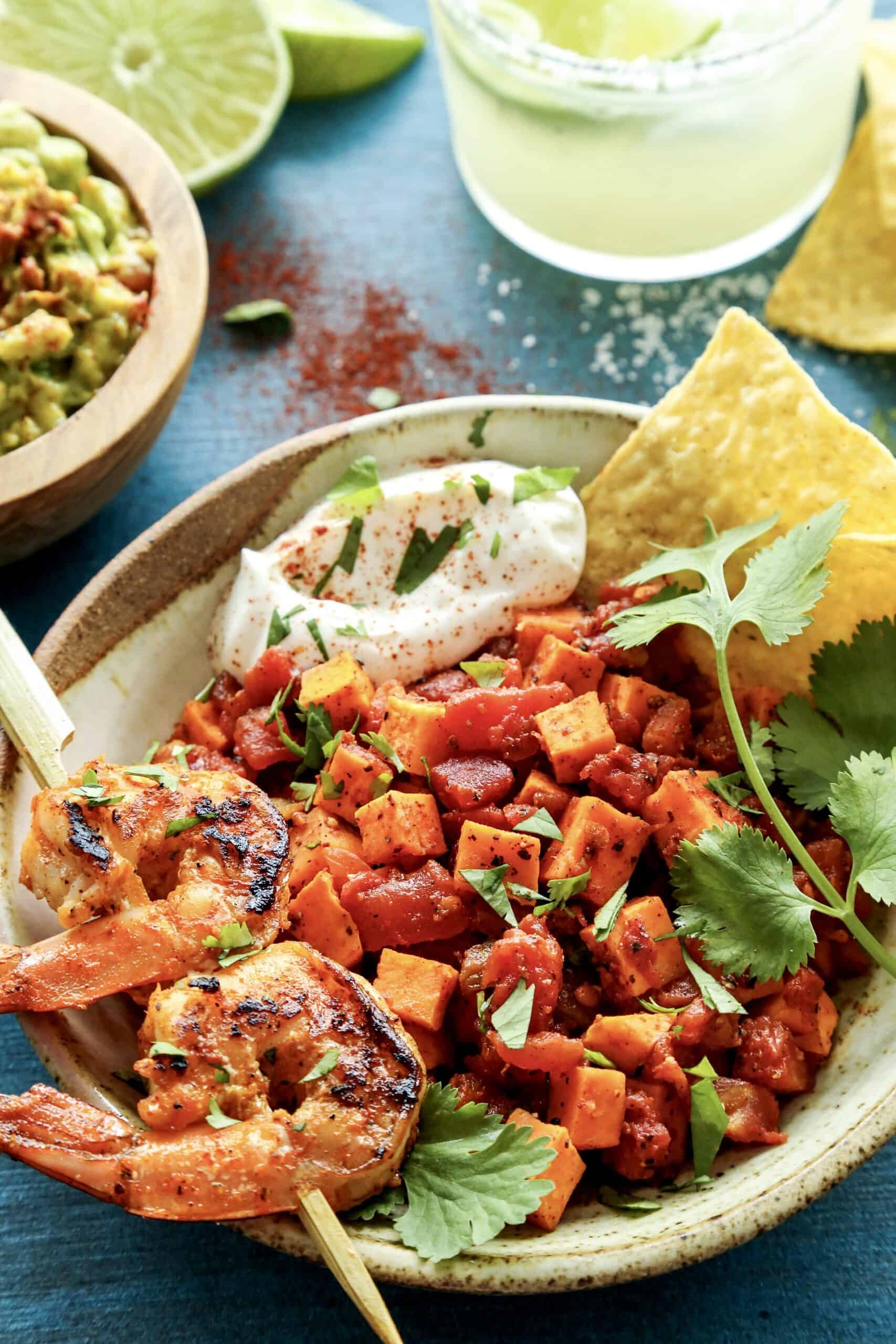Mexican Grilled Shrimp Bowls with Sweet Potato and Tomato are packed with flavor. Enjoy with chips, as a smaller appetizer or a larger meal! a skewer with 2 grilled shrimp in a bowl of diced sweet potatoes and tomatoes, plus a bowl of guacamole and a margarita