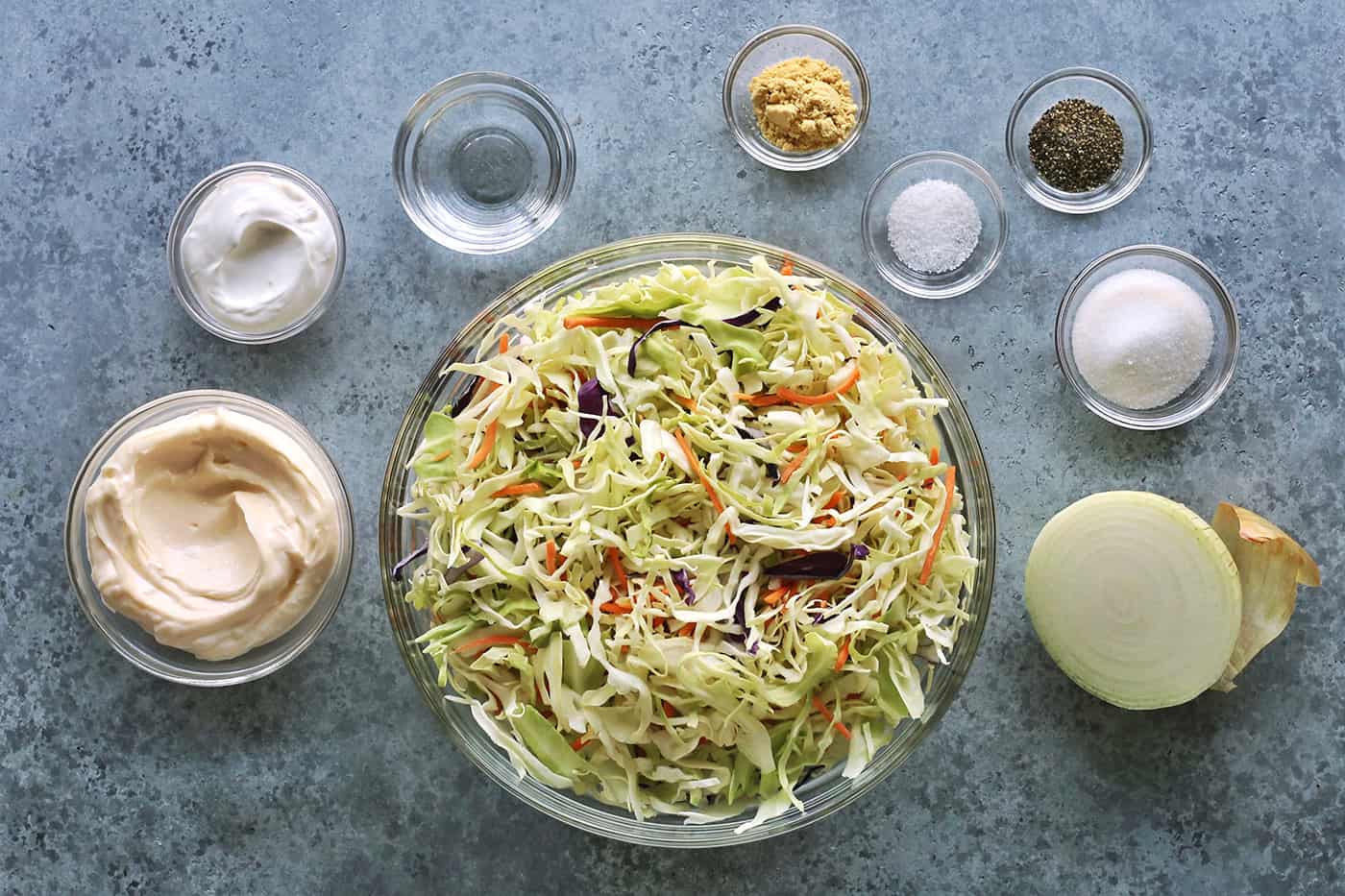 coleslaw ingredients in clear bowls: shredded cabbage, mayonnaise, sour cream, vinegar, ground mustard, salt, pepper, sugar, and half a yellow onion