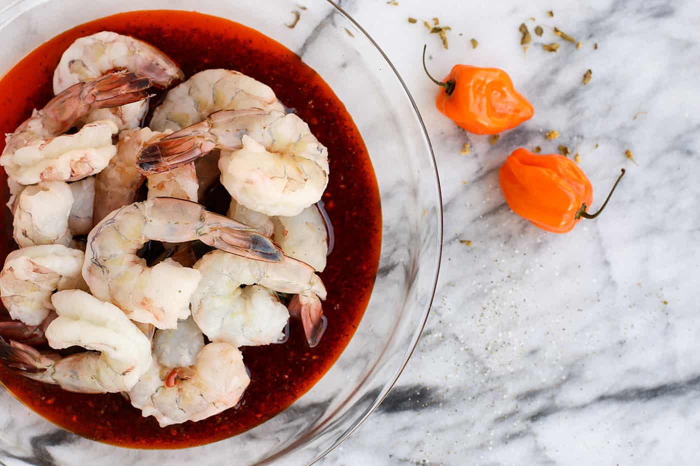 Overhead view of raw shrimp in a bowl of marinade next to two mini peppers
