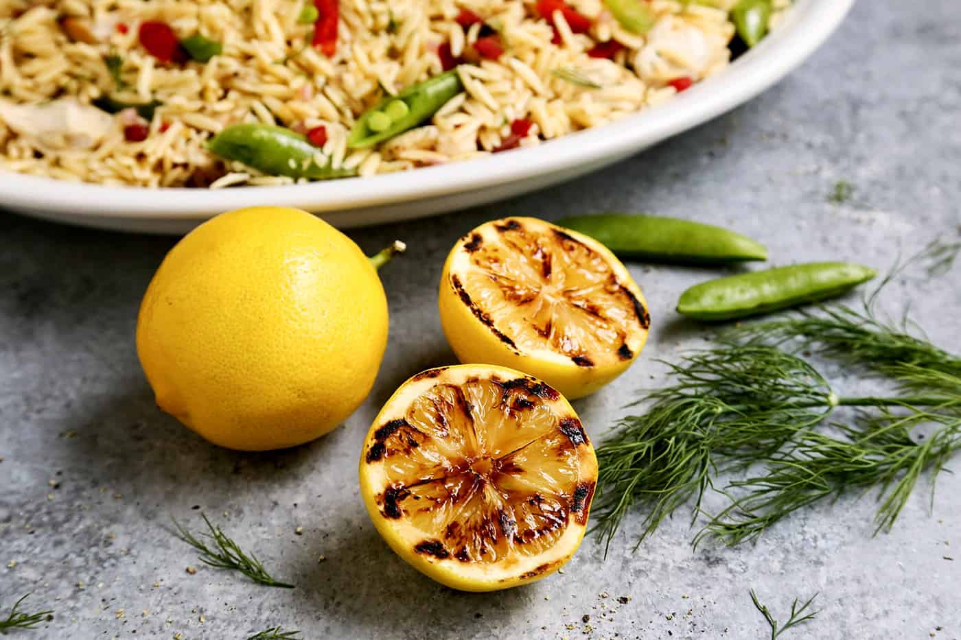 Grilled Lemon halves next to a big white bowl of Grilled Chicken and Lemon Orzo salad