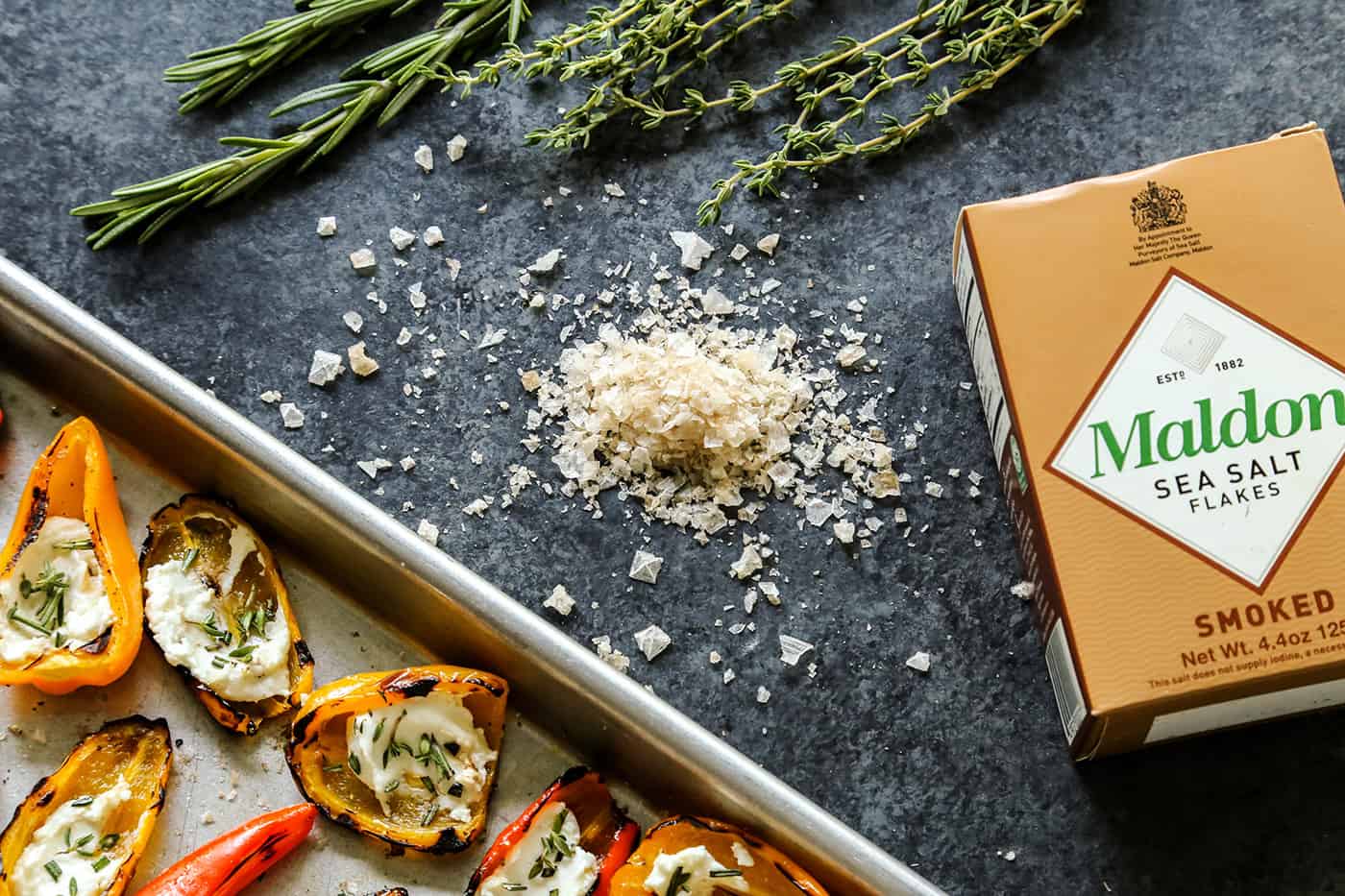 Grilled Mini Sweet Peppers with Goat Cheese on a sheet pan next to Maldon Smoked Sea Salt Flakes and a box of sea salt