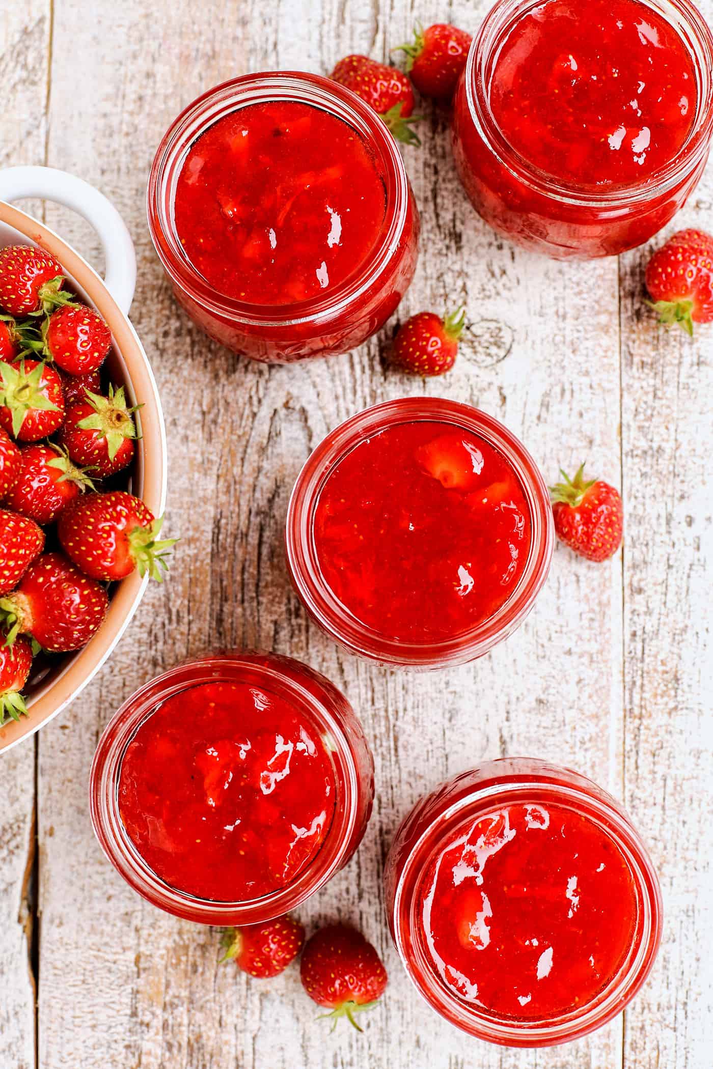 Jars of strawberry jam on a wood table next to a bowl of berries