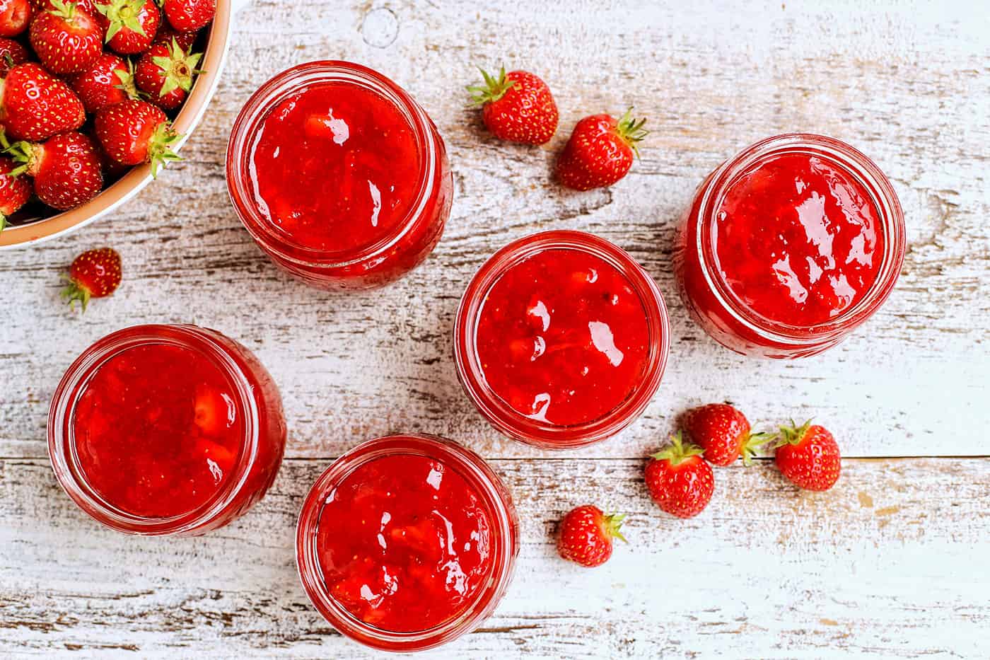 Five jars with strawberry jam next to a bowl of berries