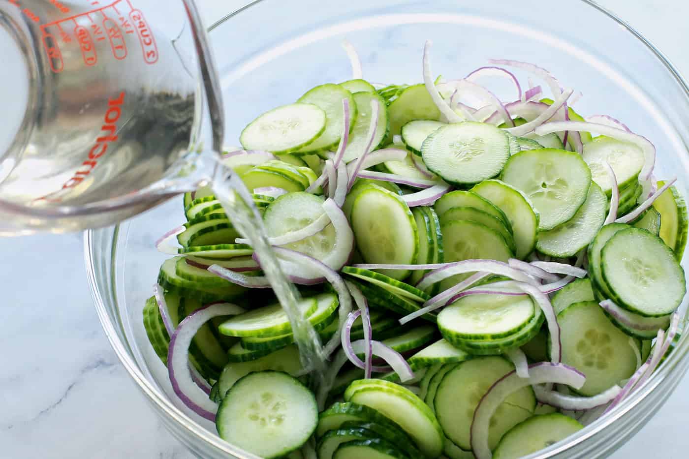 pouring vinegar mixture over sliced cucumbers and red onion in a clear bowl