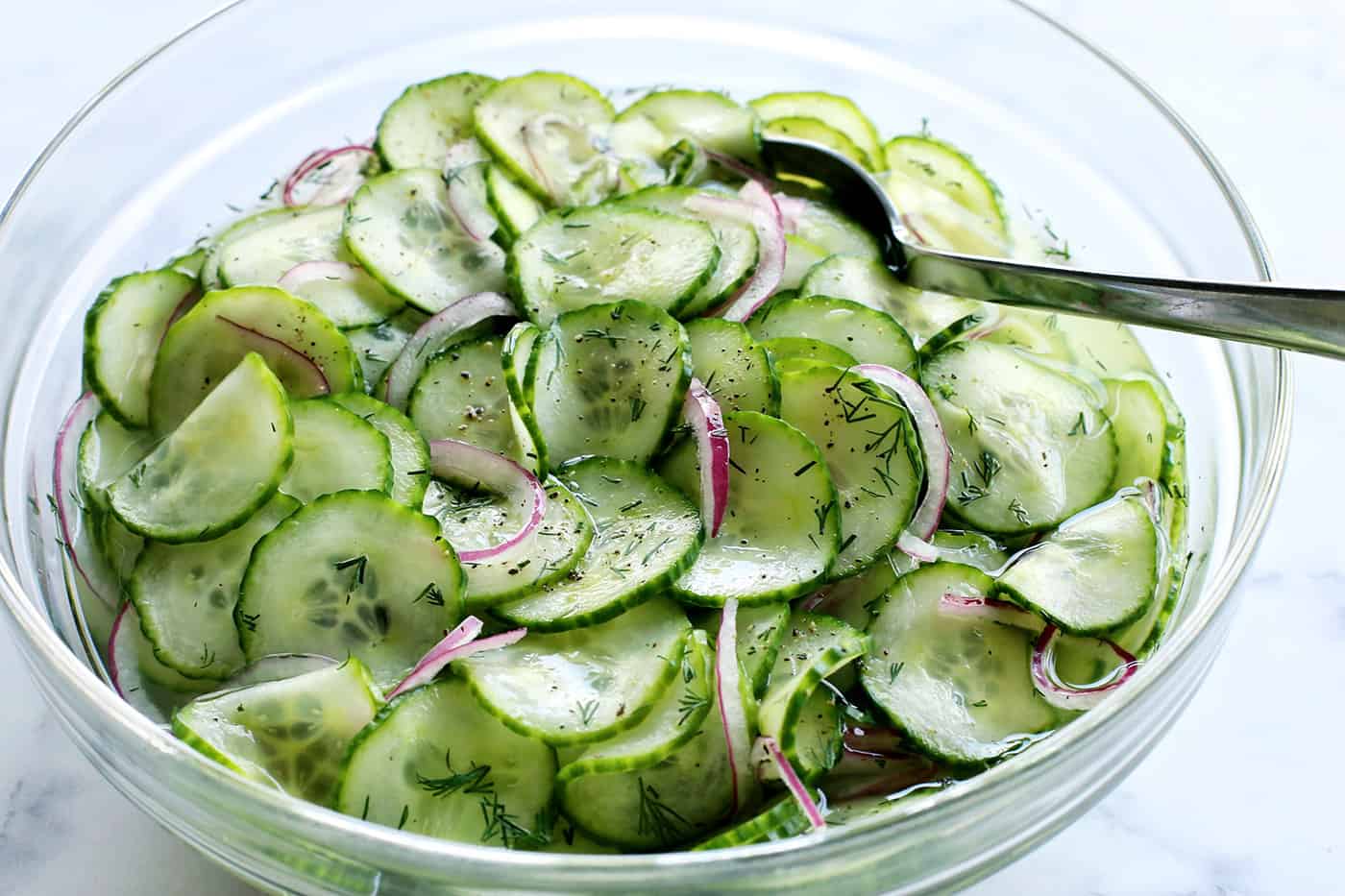 cucumber and onion salad with vinegar dressing in a large clear bowl, with a serving spoon