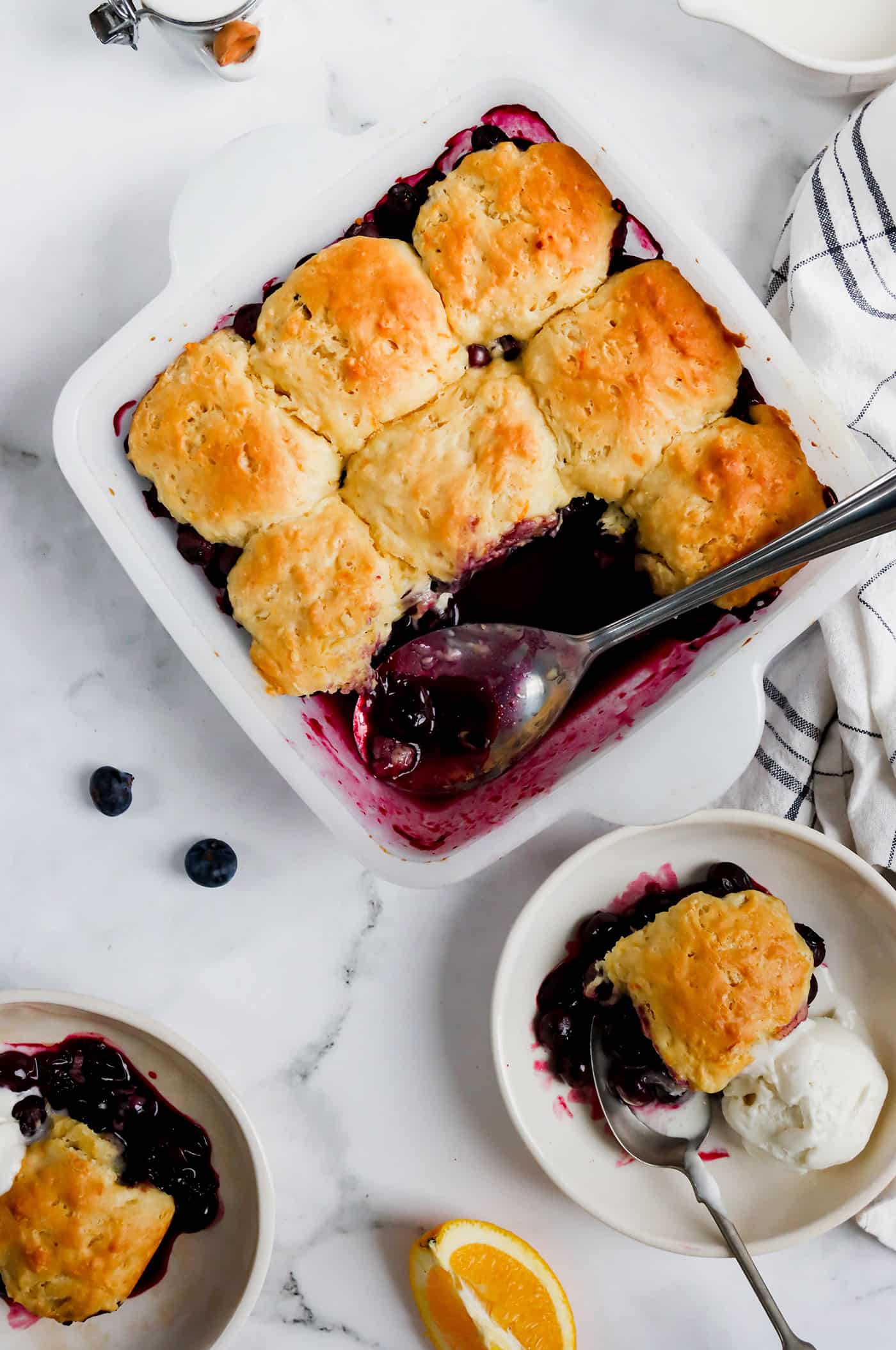 a square white pan of homemade blueberry cobbler with individual servings in small white bowls with ice cream