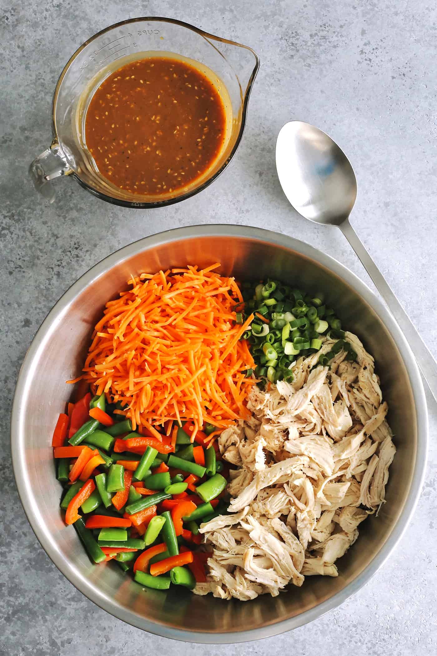 shredded chicken and cut vegetables in a large stainless steel bow, with a glass measure of peanut dressing on the side