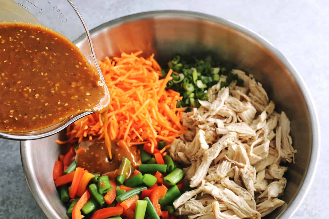 pouring creamy peanut sauce over stainless steel bowl of shredded chicken and chopped vegetables