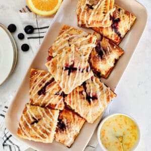 Overhead view of a platter of blueberry hand pies drizzled with orange glaze, next to small bowls with pie ingredients.