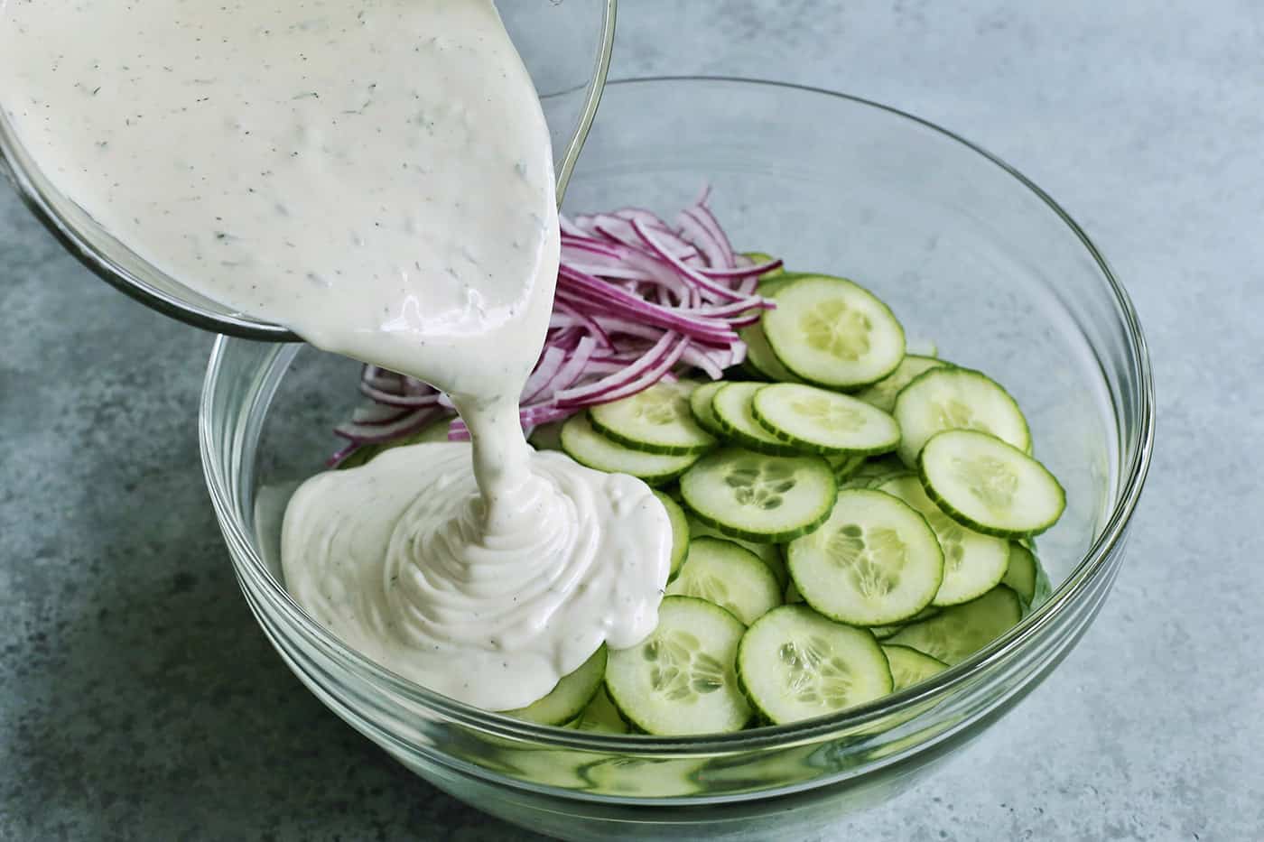 pouring creamy dressing over a bowl of sliced cucumbers and red onion