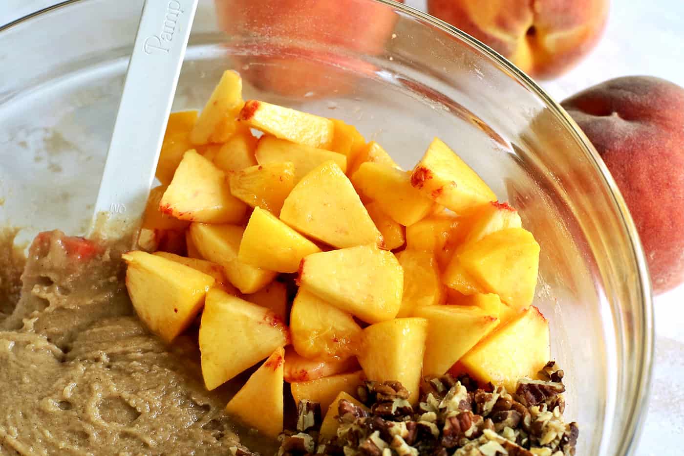 a clear mixing bowl with batter, chopped nuts, and chopped fresh peaches