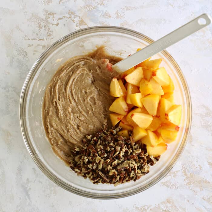 a clear mixing bowl with peach bread batter plus fresh chopped peaches and pecans