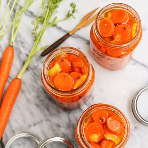 three jars of pickled carrots and a couple whole carrots with their green tops