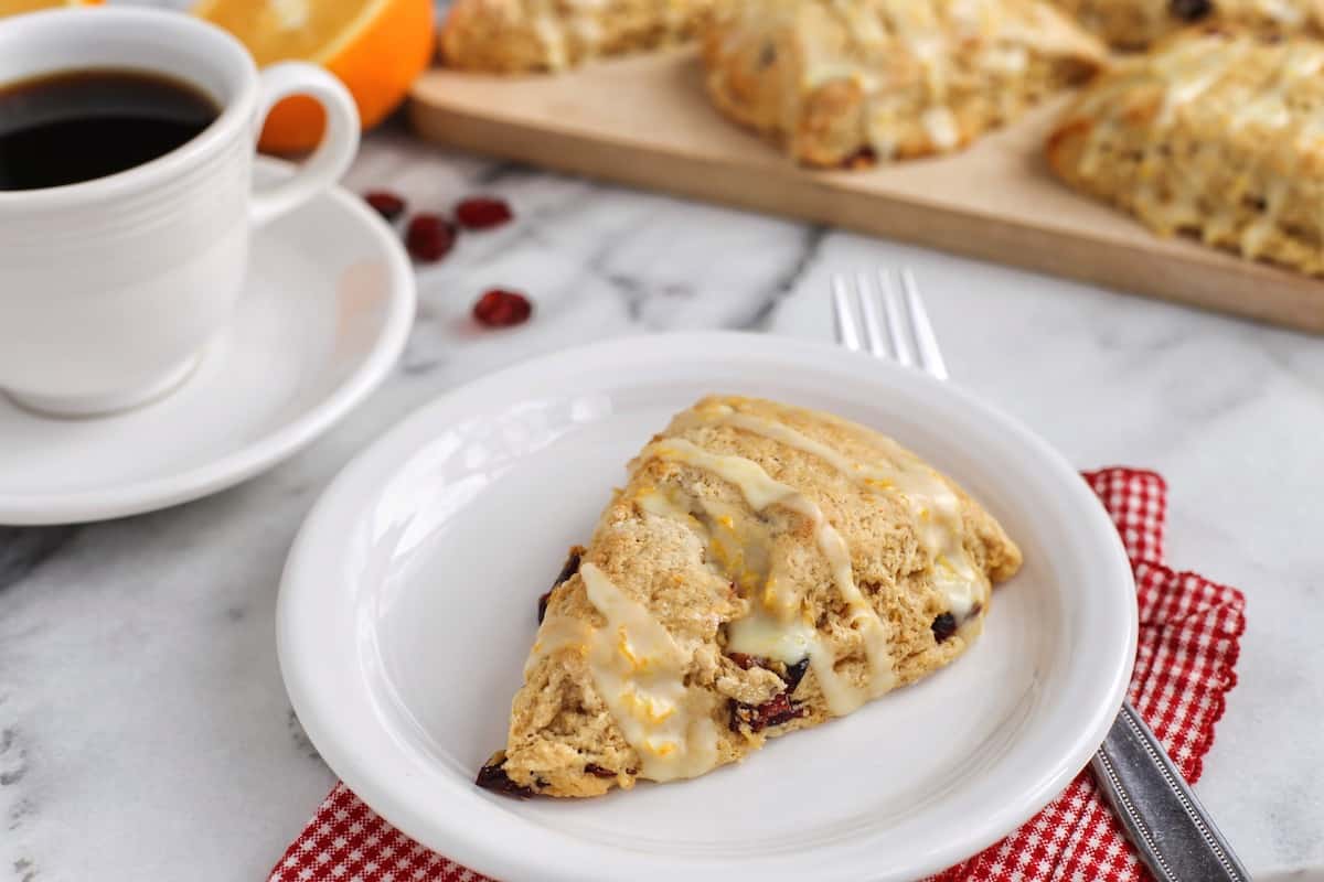 A cranberry orange scone drizzled with glaze on a white plate, with more scones on a wooden cutting board next to a cup of coffee in the background.