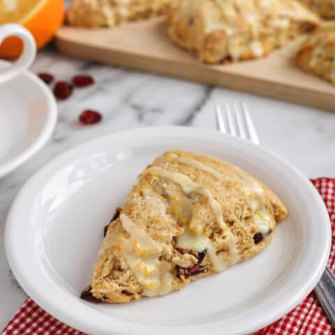 A cranberry orange scone drizzled with glaze on a white plate, with more scones on a wooden cutting board in the background.